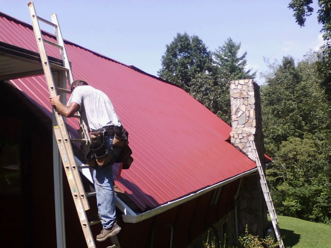 Roofer on a ladder beside a red metal roof and stone chimney — Soffit & Fascia Repair by Roofing Brothers in Yorkshire